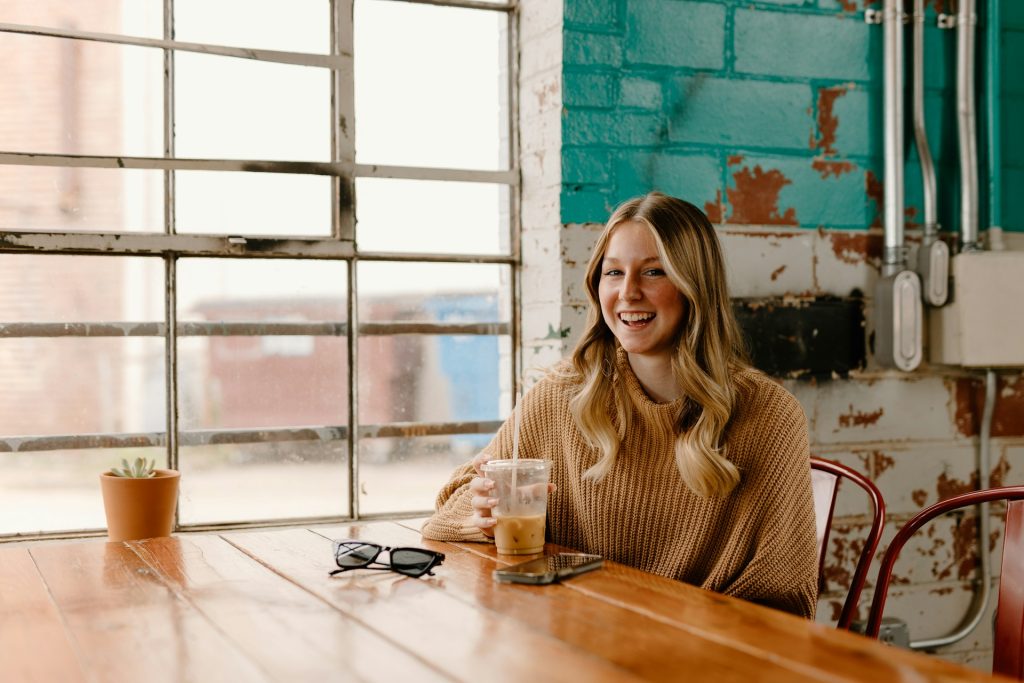 Blondi nainen istuu pöydän äärellä hymyillen. Hänellä on toisessa kädessä jääkahvi ja pöydällä on puhelin ja aurinkolasit.